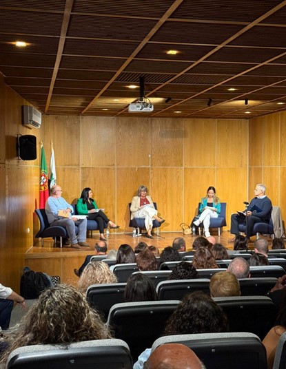 No palco, há cinco cadeiras dispostas em semicírculo, cada uma ocupada por uma pessoa, todas adultas. Da esquerda para a direita: 1. Um homem de cabelo grisalho, usando óculos, camisa azul-clara e calça azul. 2. Uma mulher com cabelo preto e liso, usando blazer verde e calças escuras. 3. Uma mulher de cabelo grisalho e liso, usa óculos, blazer bege e calça branca. 4. Outra mulher de cabelo castanho, veste blazer verde claro e calça branca. 5. Um homem de cabelo grisalho, veste suéter azul-marinho sobre camisa clara. Atrás do painel estão duas bandeiras: uma de Portugal (verde e vermelha) e outra predominantemente branca (possivelmente institucional ou regional). No teto de madeira, vê-se o projetor central, luzes embutidas e painéis de madeira alinhados. A plateia está cheia. Todos assistem atentos, indicando um ambiente de debate ou conferência formal. Não há elementos de distração ou objetos adicionais no palco, além das cadeiras e bandeiras.