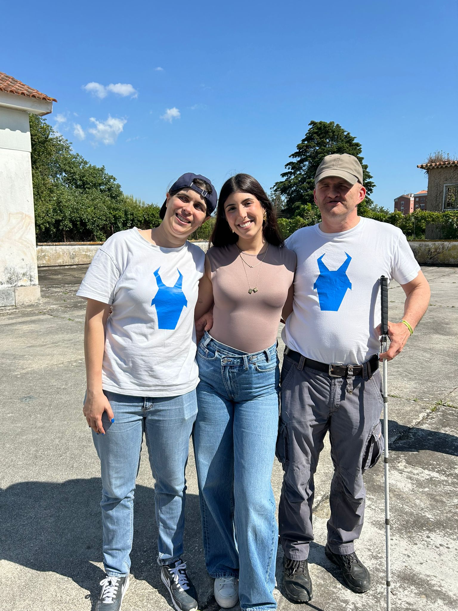 Três pessoas juntas ao ar livre num dia ensolarado, num pátio de cimento, com céu azul e algumas árvores ao fundo. À esquerda, uma mulher está a sorrir. Usa um boné escuro virado para trás, t-shirt branca da Associação Cabra Cega, calças de ganga e ténis escuros. No centro, uma mulher está a sorrir. Tem cabelo longo e escuro, usa uma blusa justa bege, calças de ganga de cintura alta e ténis claros. Ela está a abraçar as outras duas pessoas, uma de cada lado. À direita, um homem usa boné claro, t-shirt branca da Associação Cabra Cega, calças cinzentas e botas escuras. Ele está a segurar uma bengala longa branca e preta na mão direita.
