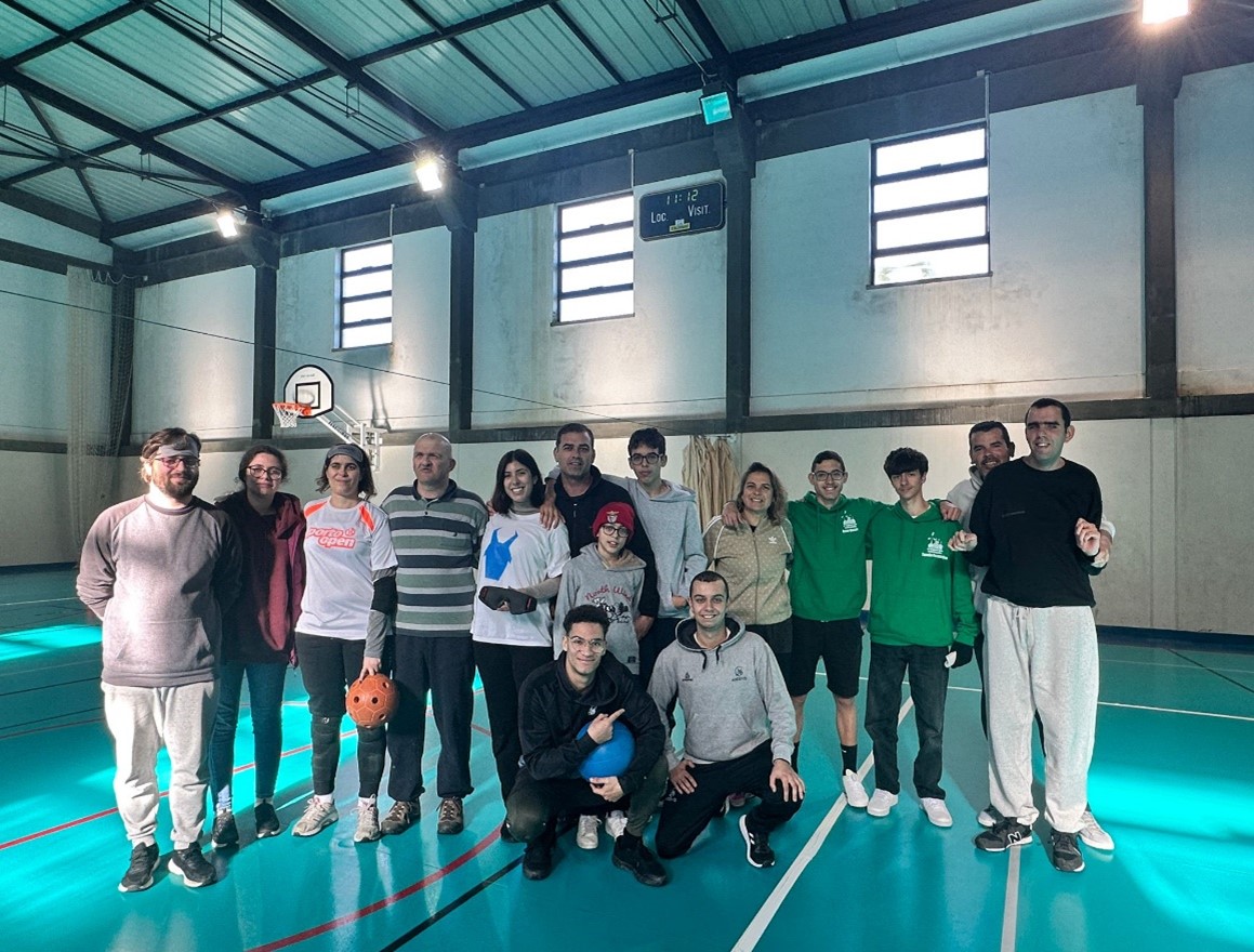 Grupo de 15 pessoas num pavilhão desportivo, iluminado por luz natural que entra pelas janelas altas e por luzes no teto. Algumas pessoas estão de pé, enquanto duas estão agachadas à frente, a sorrir para a câmara. Uma mulher segura uma bola laranja de Goalball. Algumas pessoas estão vestidas com roupas de desporto.
