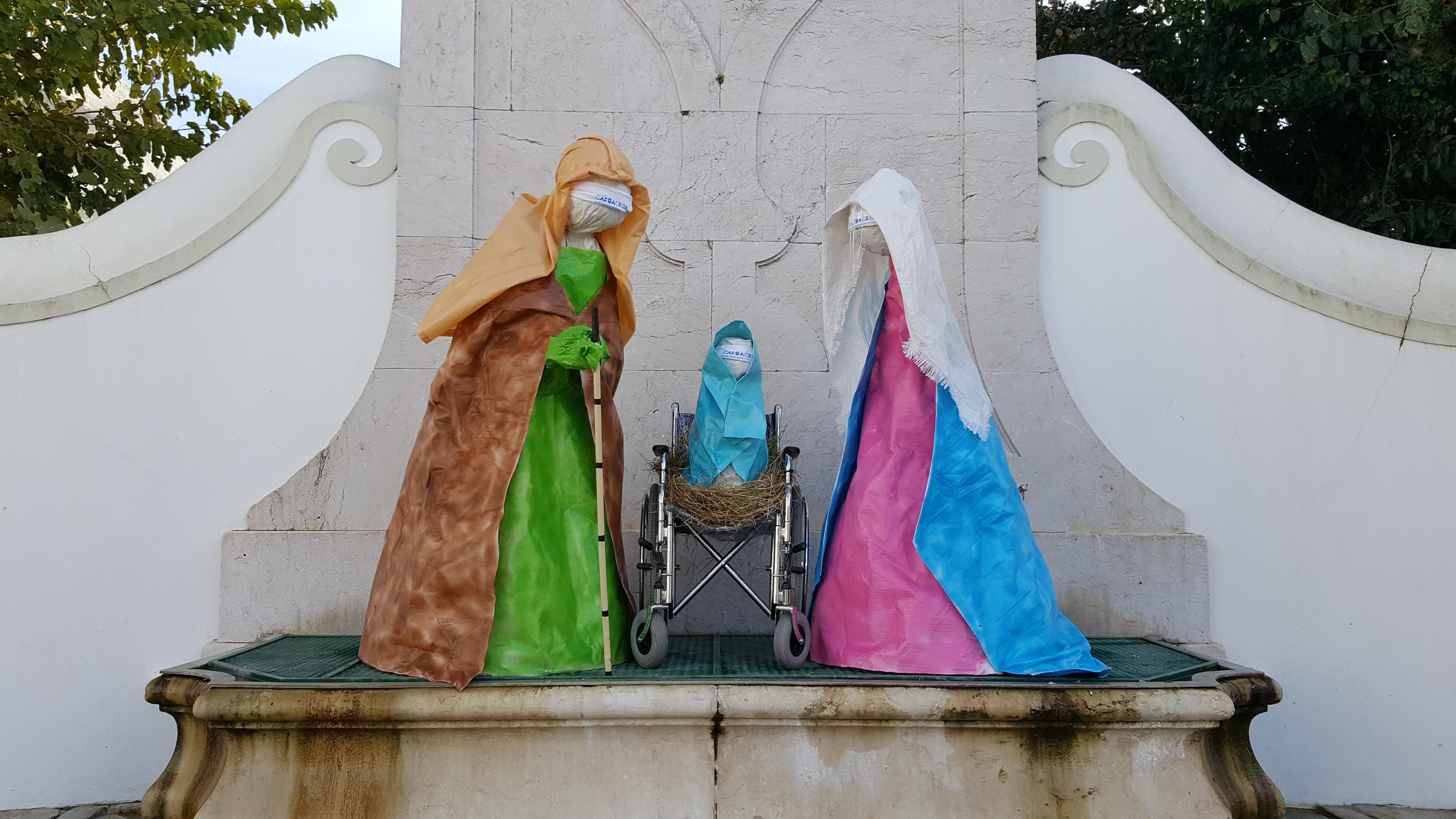 Foto do Chafariz pombalino de Sobral onde está montado o presépio da Cabra Cega sob o tanque central e à direita duas faces da estrutura identificativa da Associação e do presépio.